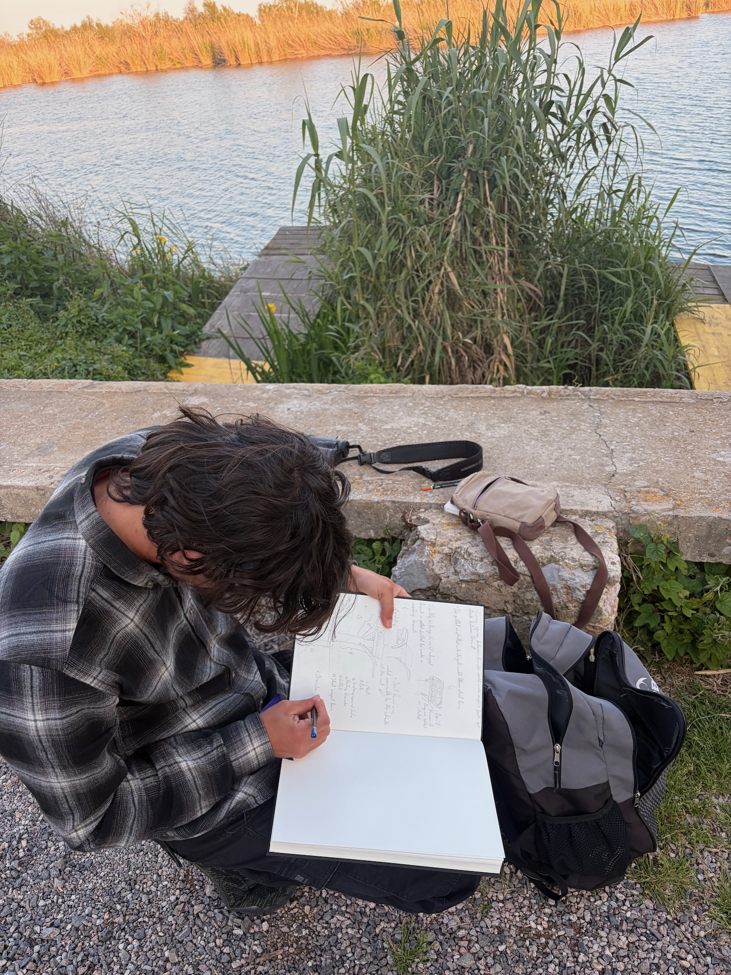 A person wearing a black and white checked shirt sits on a stone wall by the water, sketching in a notebook. A black bag is beside them, with tall grass and trees visible in the background.