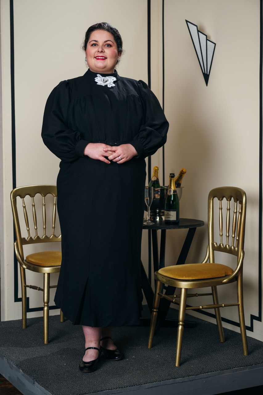 A woman in a long black dress with puffed sleeves stands in front of a small table featuring champagne bottles. Two gold chairs are beside her, and the backdrop consists of a plain wall with a decorative geometric design.