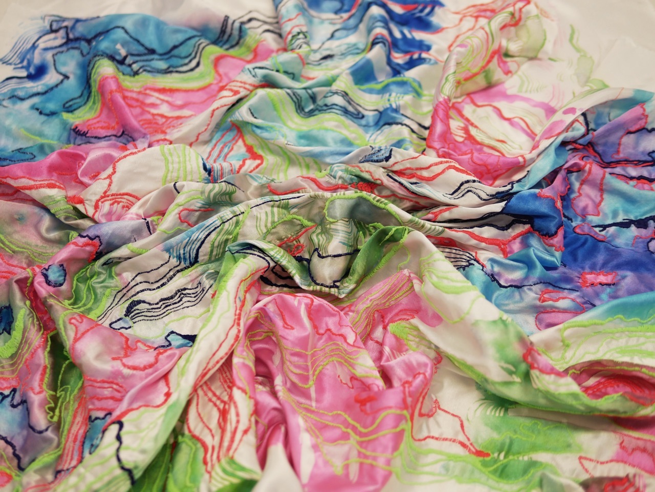 Crumpled fabric with flowing watercolour patterns in pink, green, blue and yellow tones creating organic curved lines and soft colour blends.
