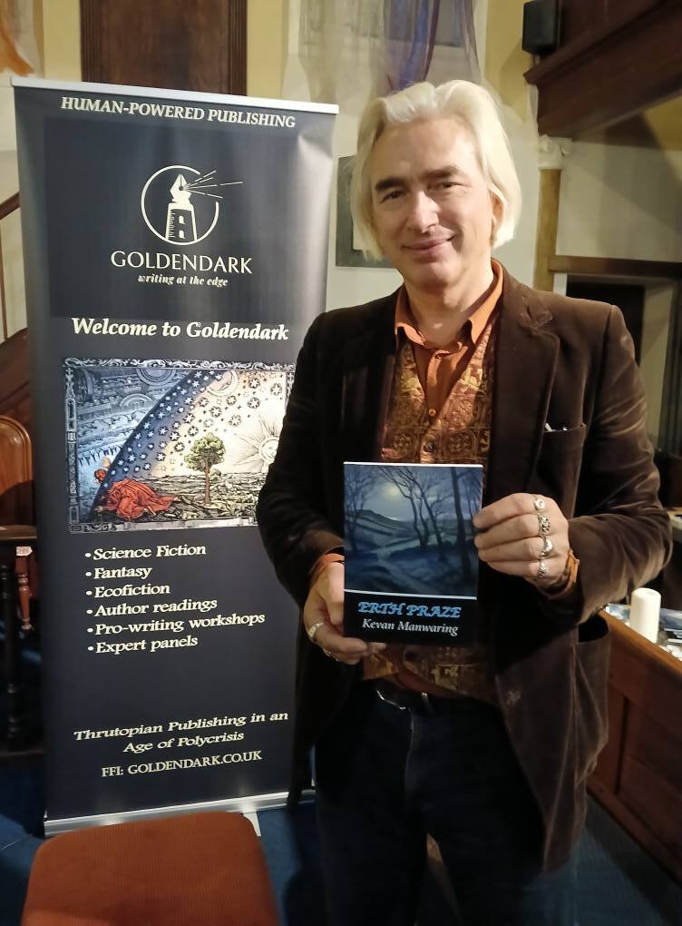 Dr K Manwaring holding copy of poetry collection "Erth Praze", standing beside black Goldendark publishing banner