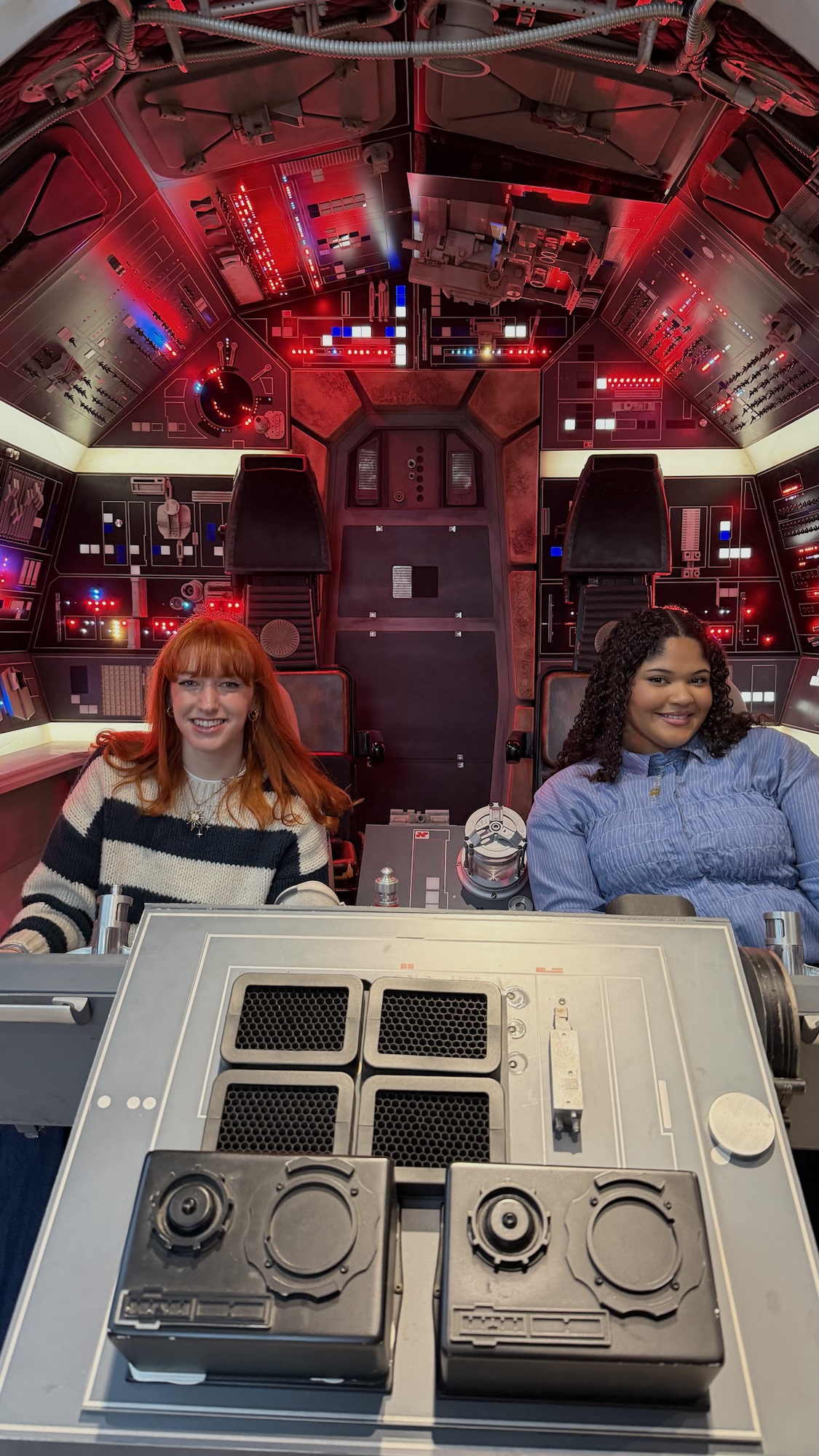 Two women sit in the cockpit of a spacecraft or simulator, with illuminated control panels and a hatchway behind them.