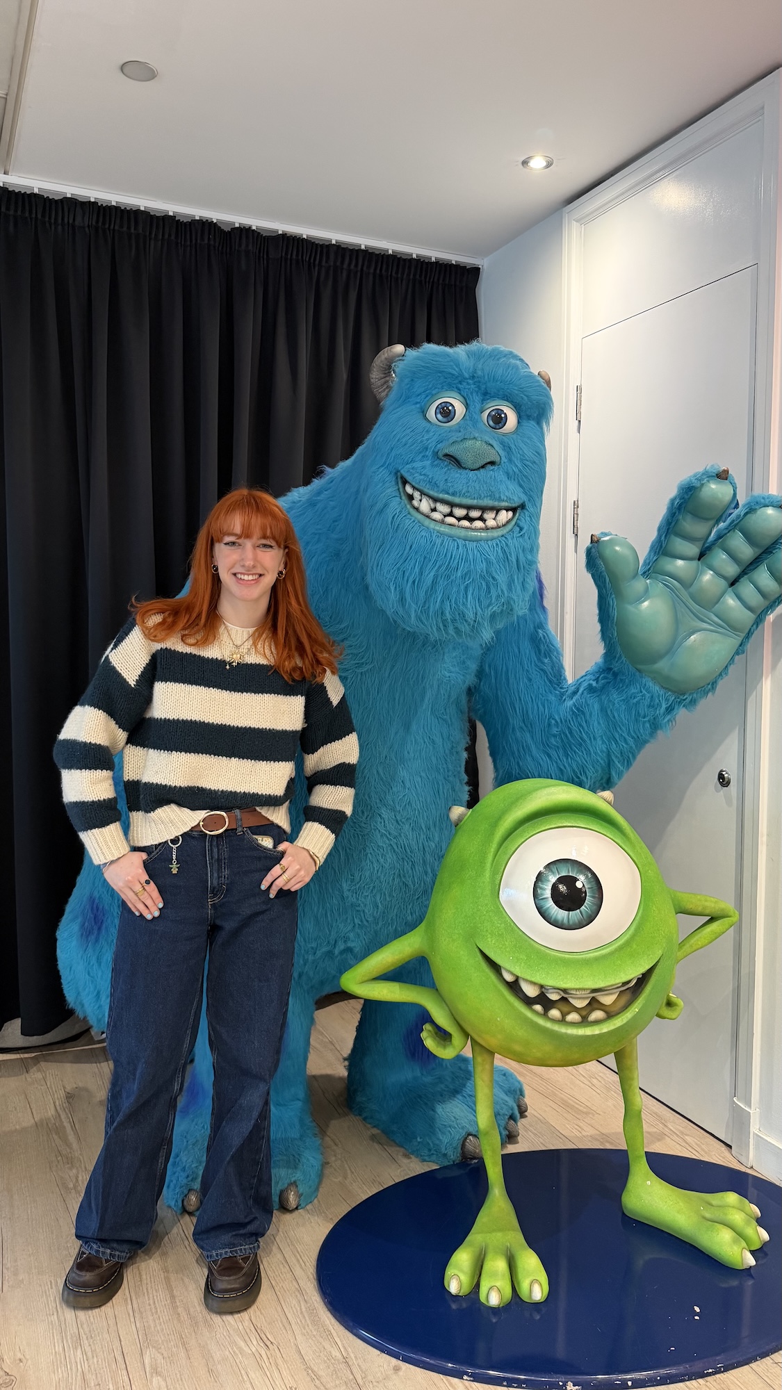 E Jarvis stands beside costumed characters Sulley and Mike Wazowski, with dark curtains and a white wall in the background.