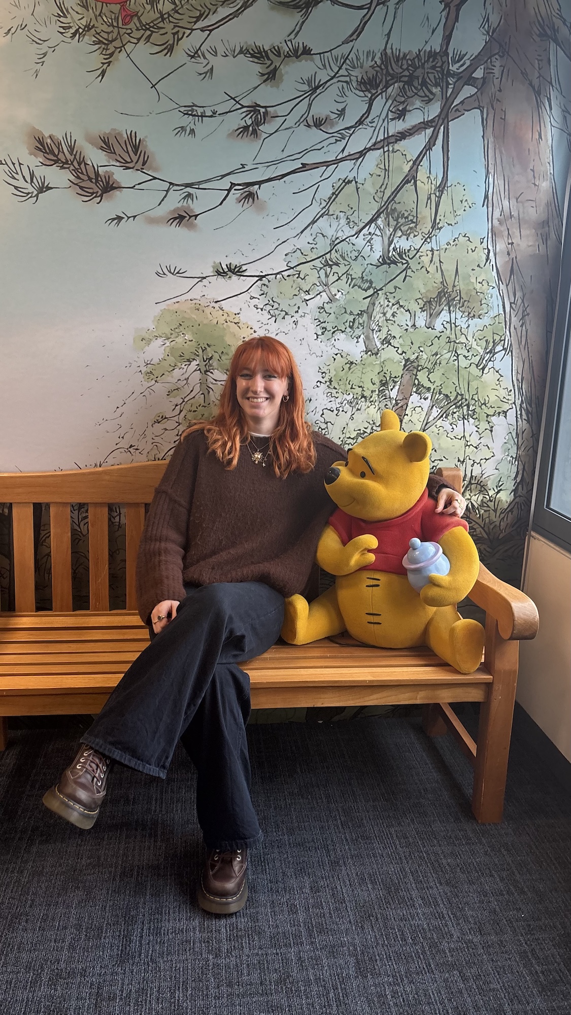 A woman with red hair sits on a wooden bench beside a Winnie the Pooh plush toy, with a painted tree mural and window in the background.