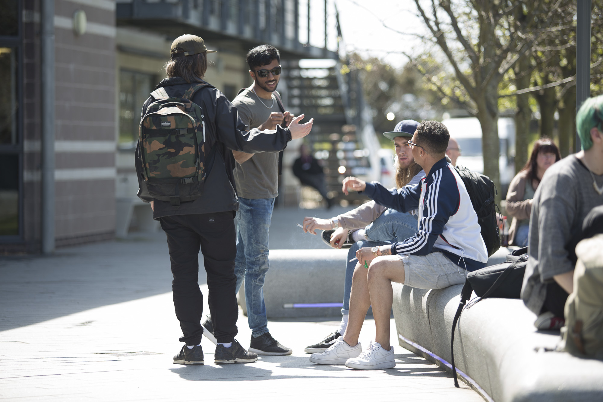 A group of students, two standing, two seated on benches, talking in a courtyard outside a university building.