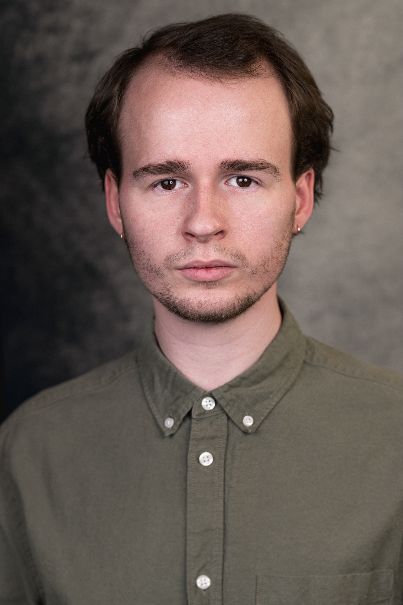 Young man with brown hair and facial hair wearing olive green button-down shirt with white buttons against grey background.