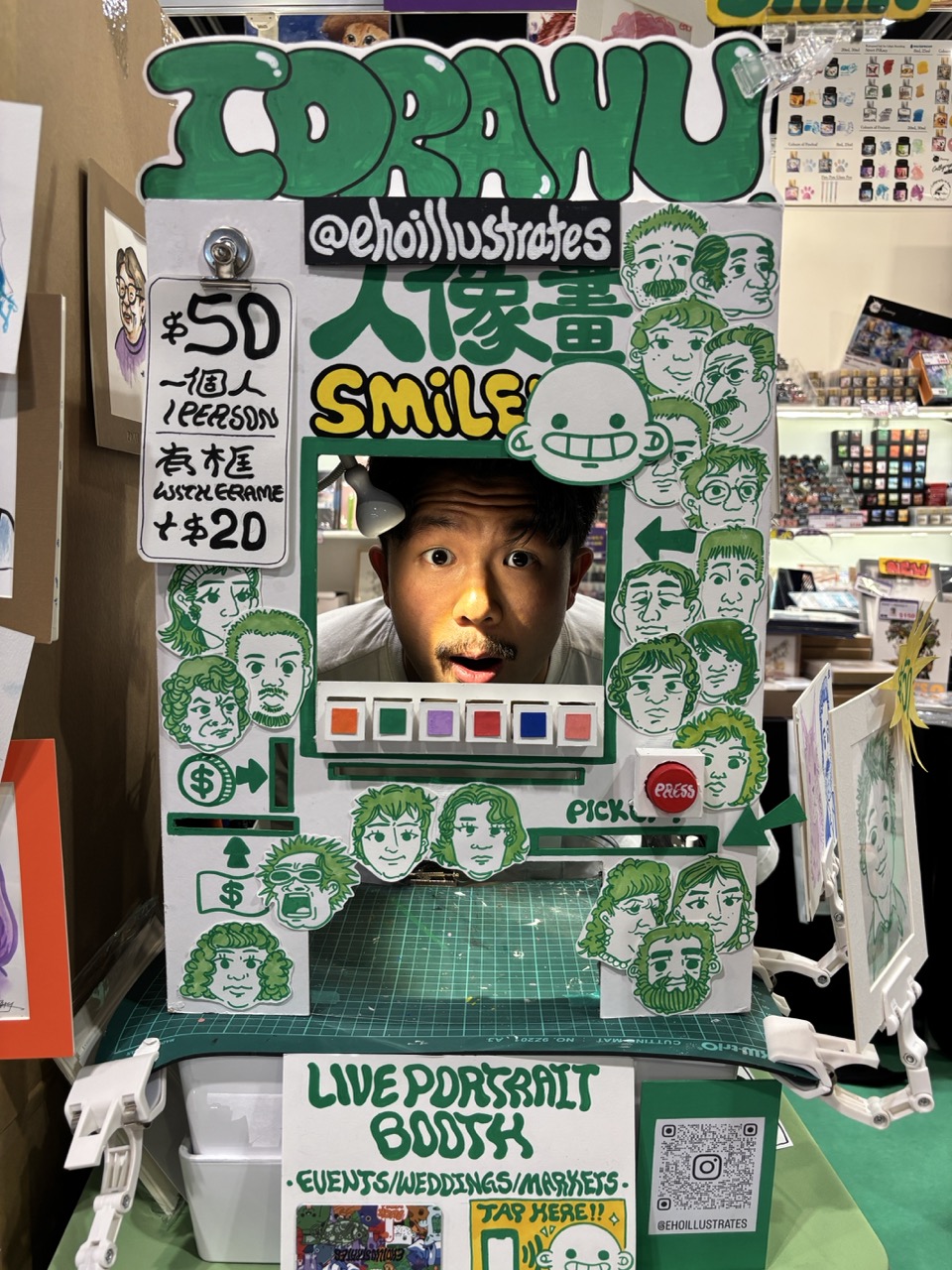A green hand-drawn portrait booth frame with the word “SMILE”, a cut-out opening for a face, and pricing signs, set on a table with art supplies and a QR code display.