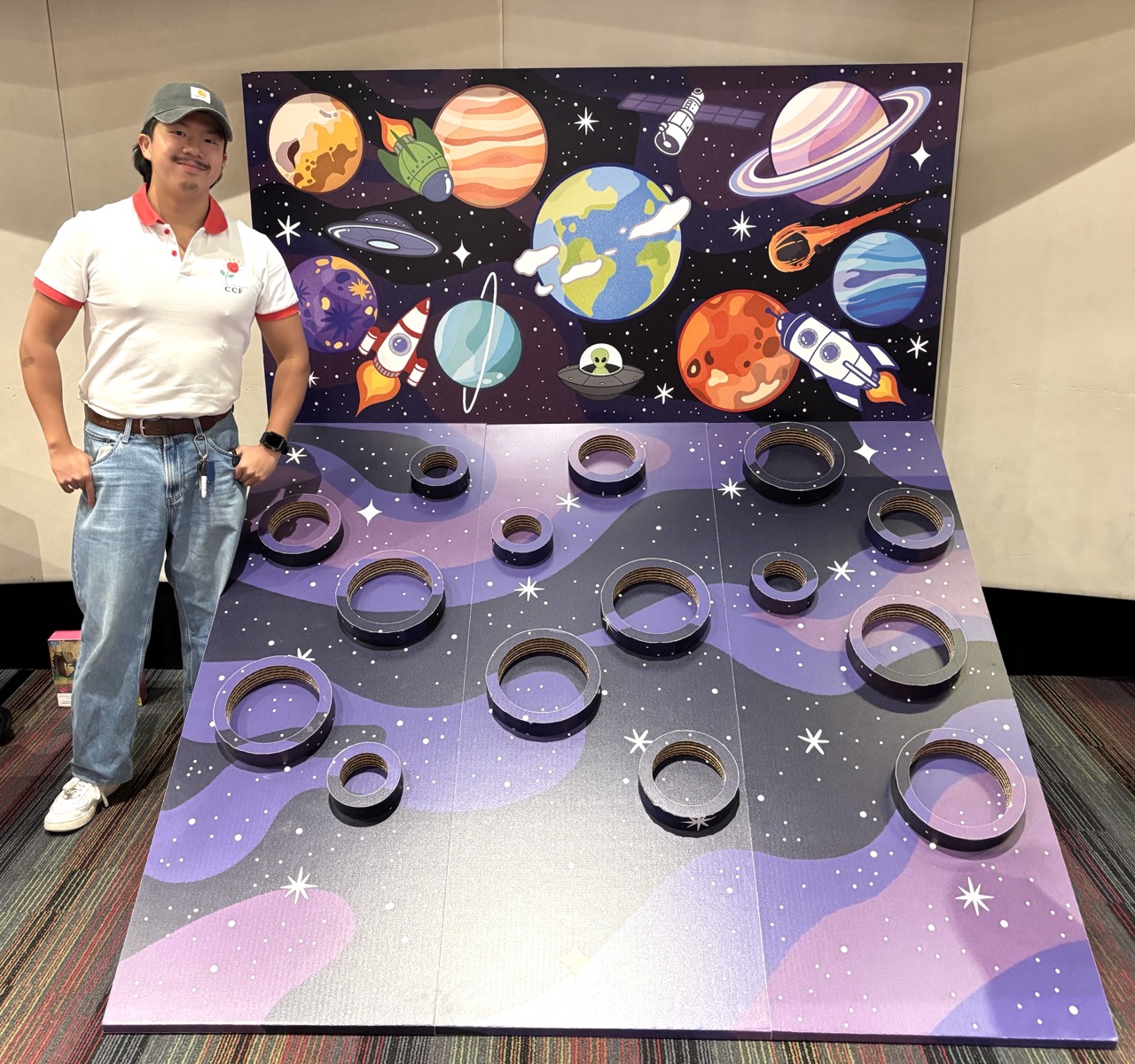 A man in a white polo shirt and blue jeans stands beside a space-themed carnival-style game board with ring targets, set against a backdrop of illustrated planets and rockets.