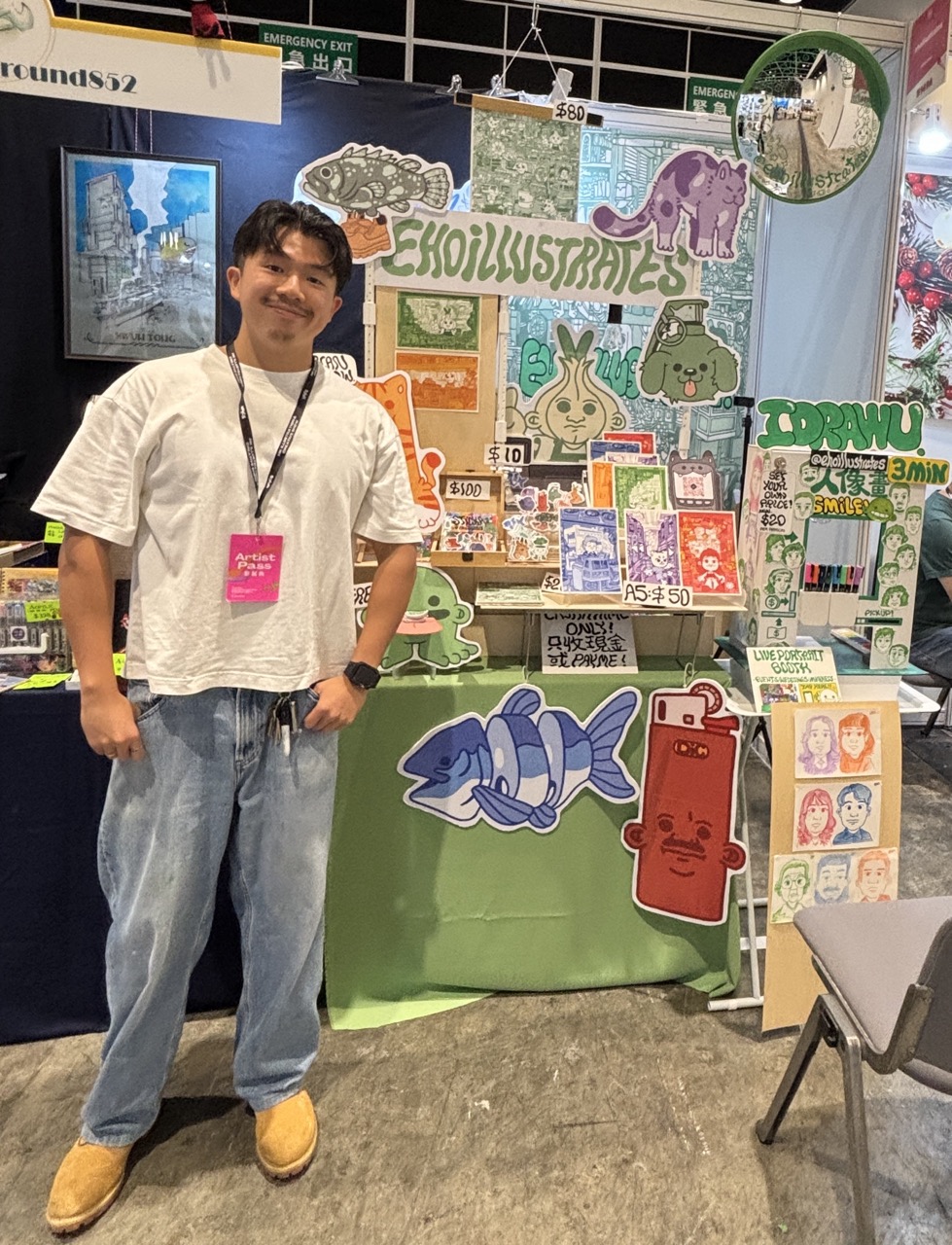 A man with a lanyard stands beside a display of illustrated prints and stickers at an indoor market stall.
