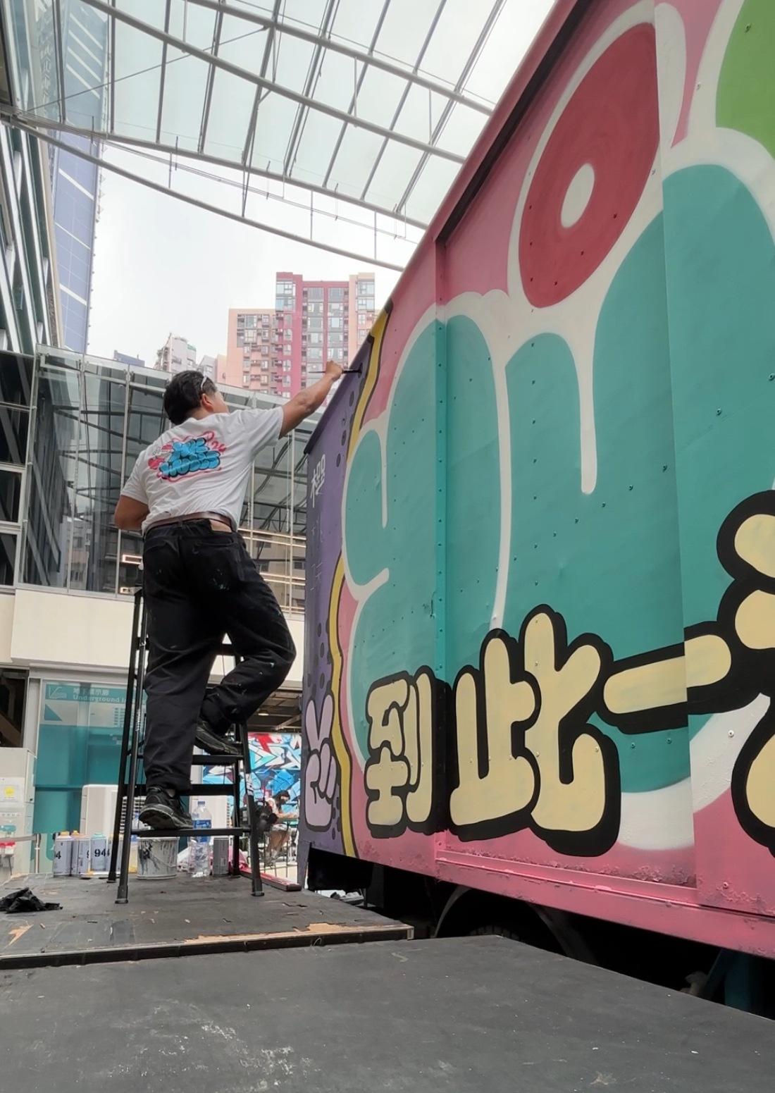 A man standing on a platform ladder paints a colourful mural on the side of a truck, with modern glass buildings and a covered walkway in the background.