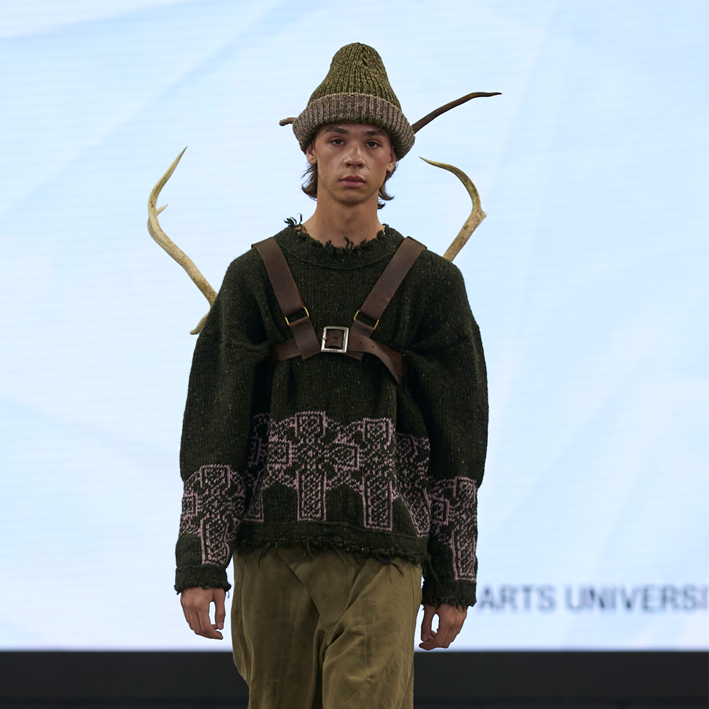 Male model on runway wearing olive green knit hat, patterned jumper, brown leather harness, and beige trousers with horned headpiece.