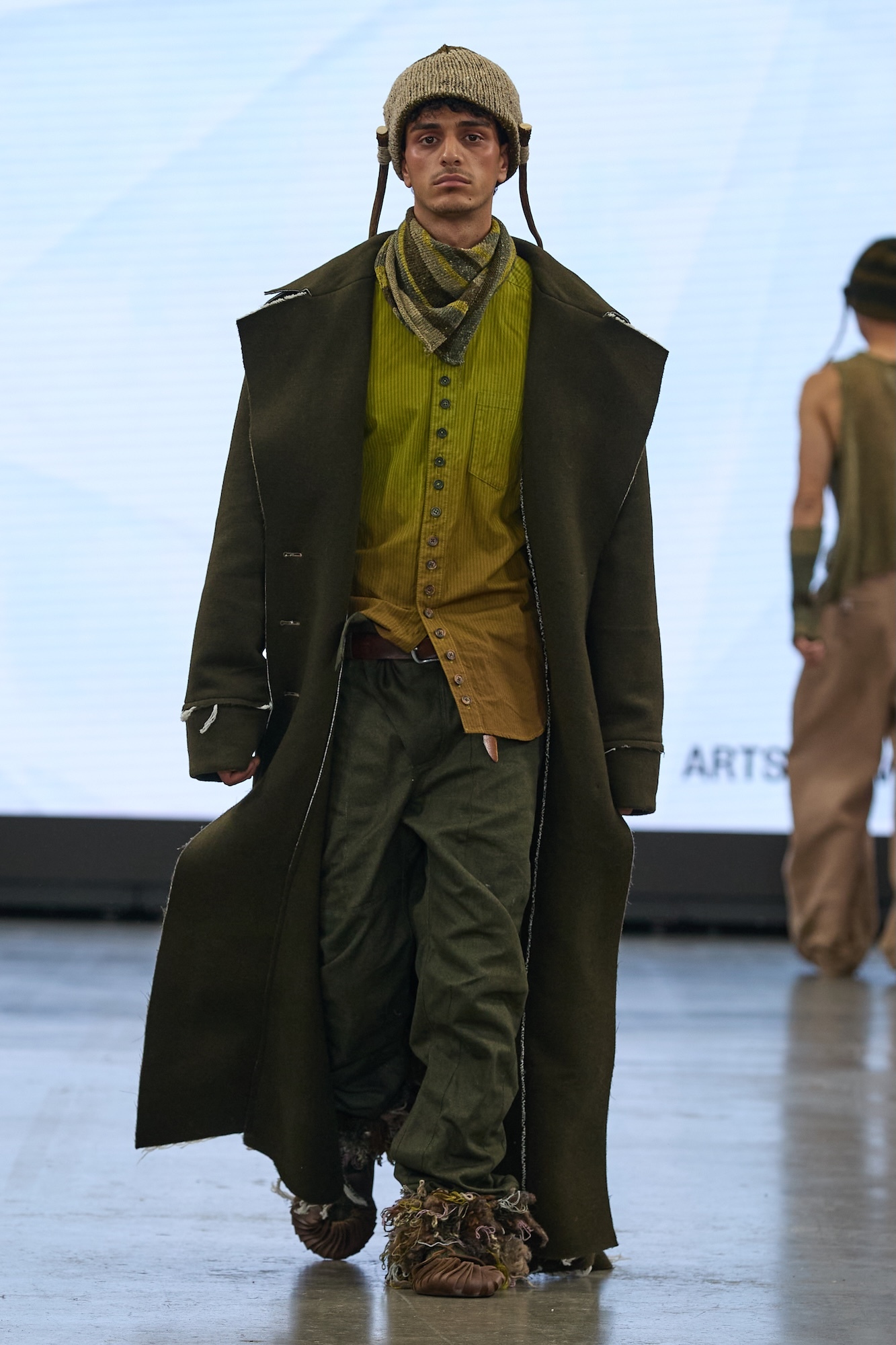 Male model on runway wearing olive green long coat, mustard waistcoat, dark trousers, knit hat with ear flaps, and textured boots.