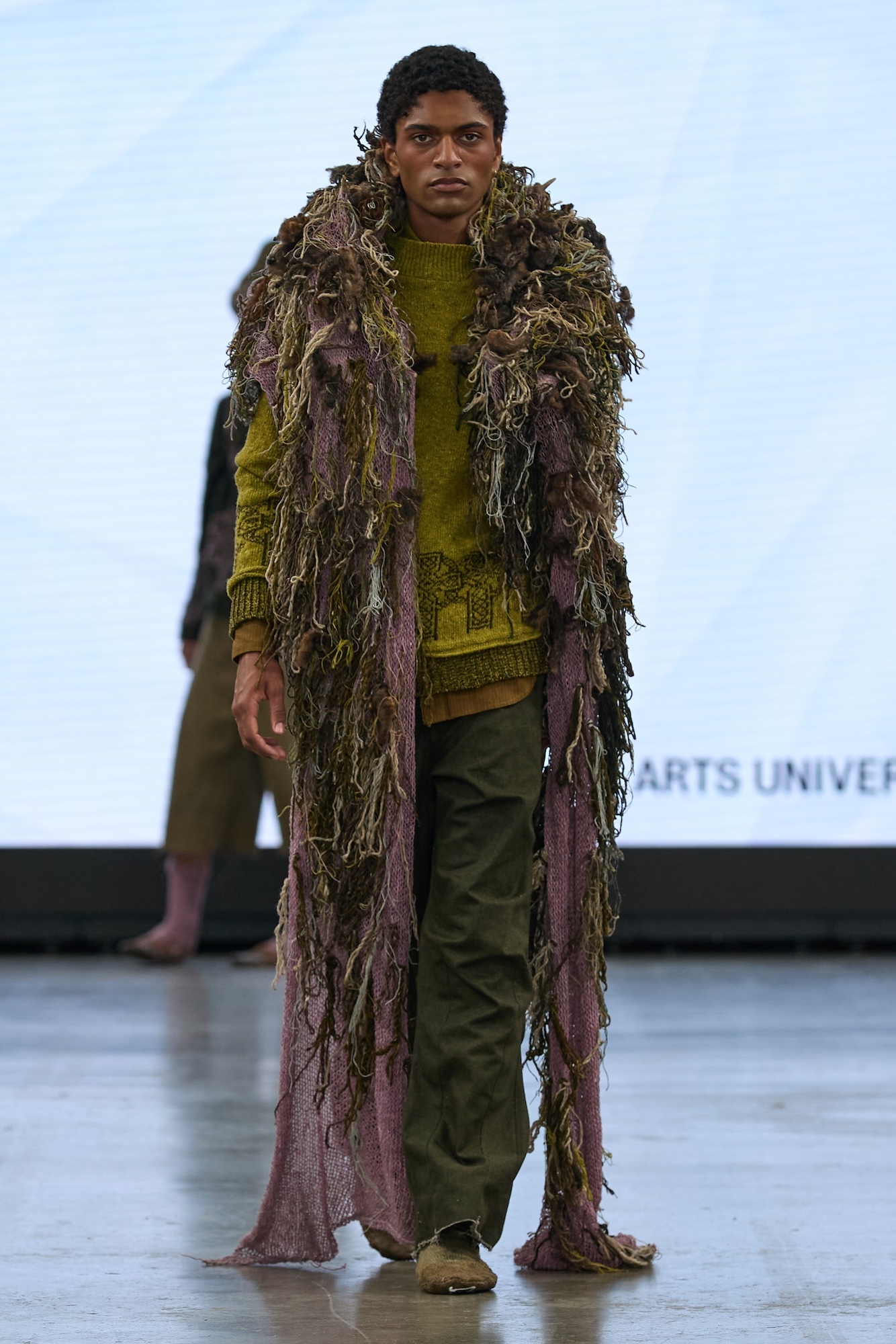Man wearing green textured jumper and trousers with brown fringe coat made of grass-like strands on white runway stage.