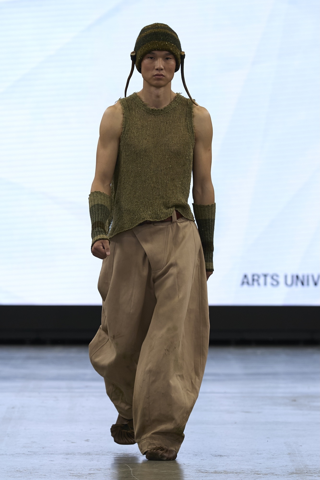 Male model on runway wearing olive green knitted vest, matching helmet with ear flaps, arm warmers, and beige wide-leg trousers.