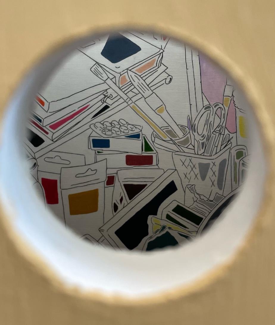 A close-up view through a circular hole, revealing a colourful assortment of art supplies, including paints, brushes, and scissors, arranged in a messy composition against a light backdrop.