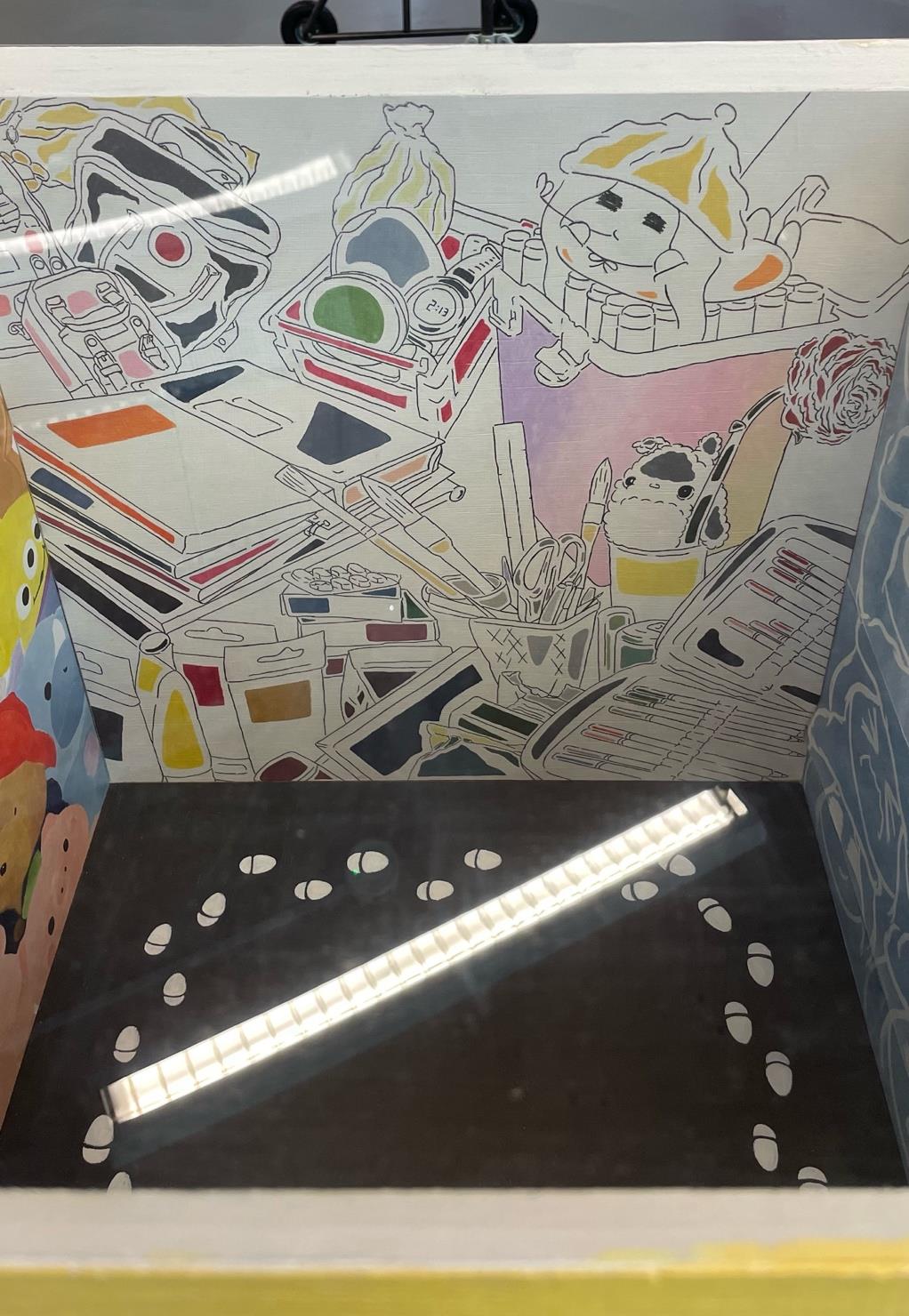 A clear display box contains a black base with white footprints surrounding it. The background features a colourful mural of various objects, including books, paints, and a cartoonish character, outlined in black and partially coloured.