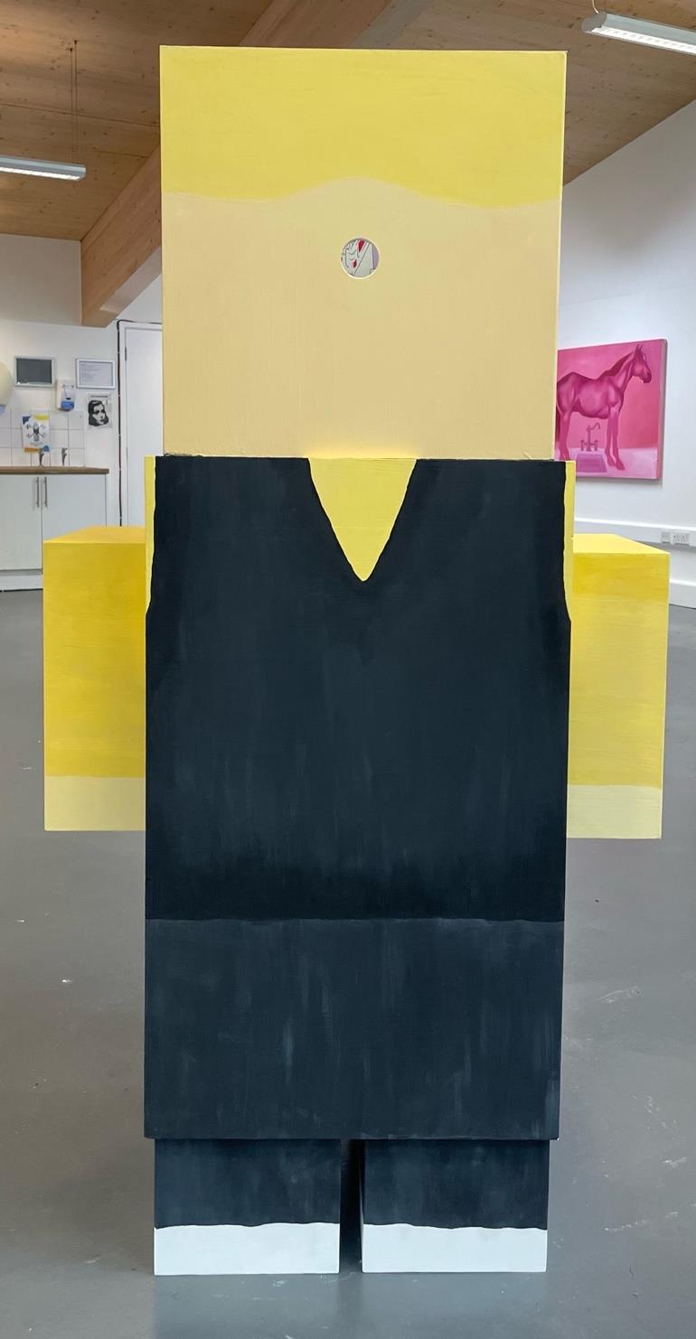 A large, three-dimensional figure made of yellow and black cardboard, resembling a blocky humanoid shape. The figure has a yellow head with a round eye, and a black sleeveless top paired with trousers, set against a minimalist gallery background.