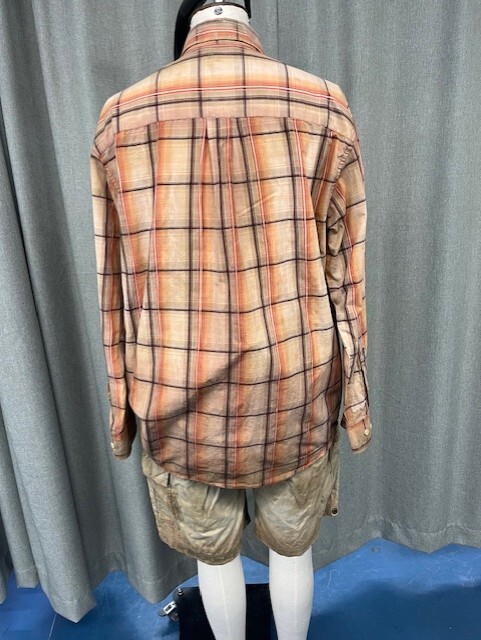 Man from behind wearing beige plaid shirt and khaki shorts with white socks against grey curtain backdrop.