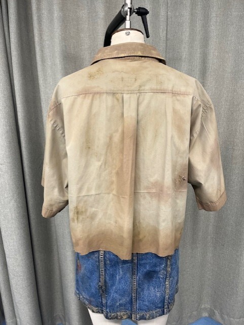 Back view of beige shirt with dark collar on mannequin, blue denim skirt below, grey curtain background.