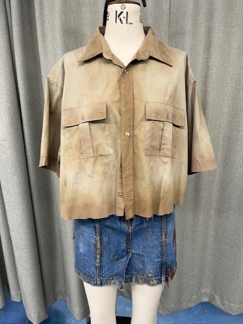 Beige short-sleeved shirt with chest pockets and blue denim skirt displayed on white mannequin against grey curtain backdrop.