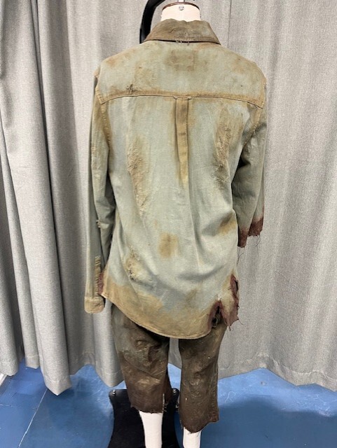 Mannequin wearing weathered olive green military uniform with brown stains, standing against grey curtain backdrop on blue floor.