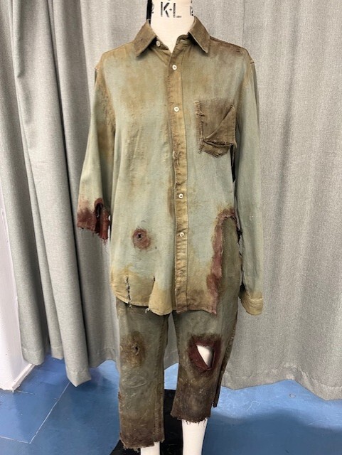 Distressed khaki shirt and trousers with brown stains and tears, displayed on mannequin against grey curtain backdrop.