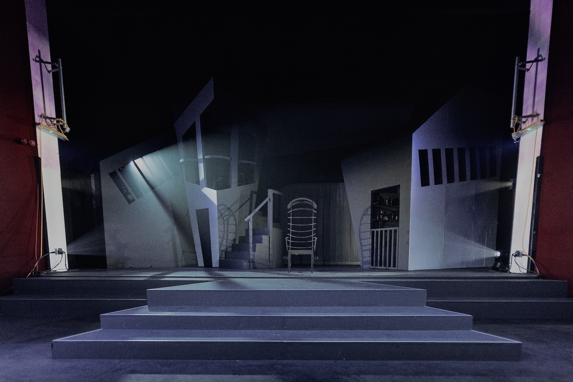 Stage set at the Palace Court Theatre, Bournemouth, with grey walls, metal staircase, and turquoise lighting beam cutting diagonally across dark space with stepped platform.