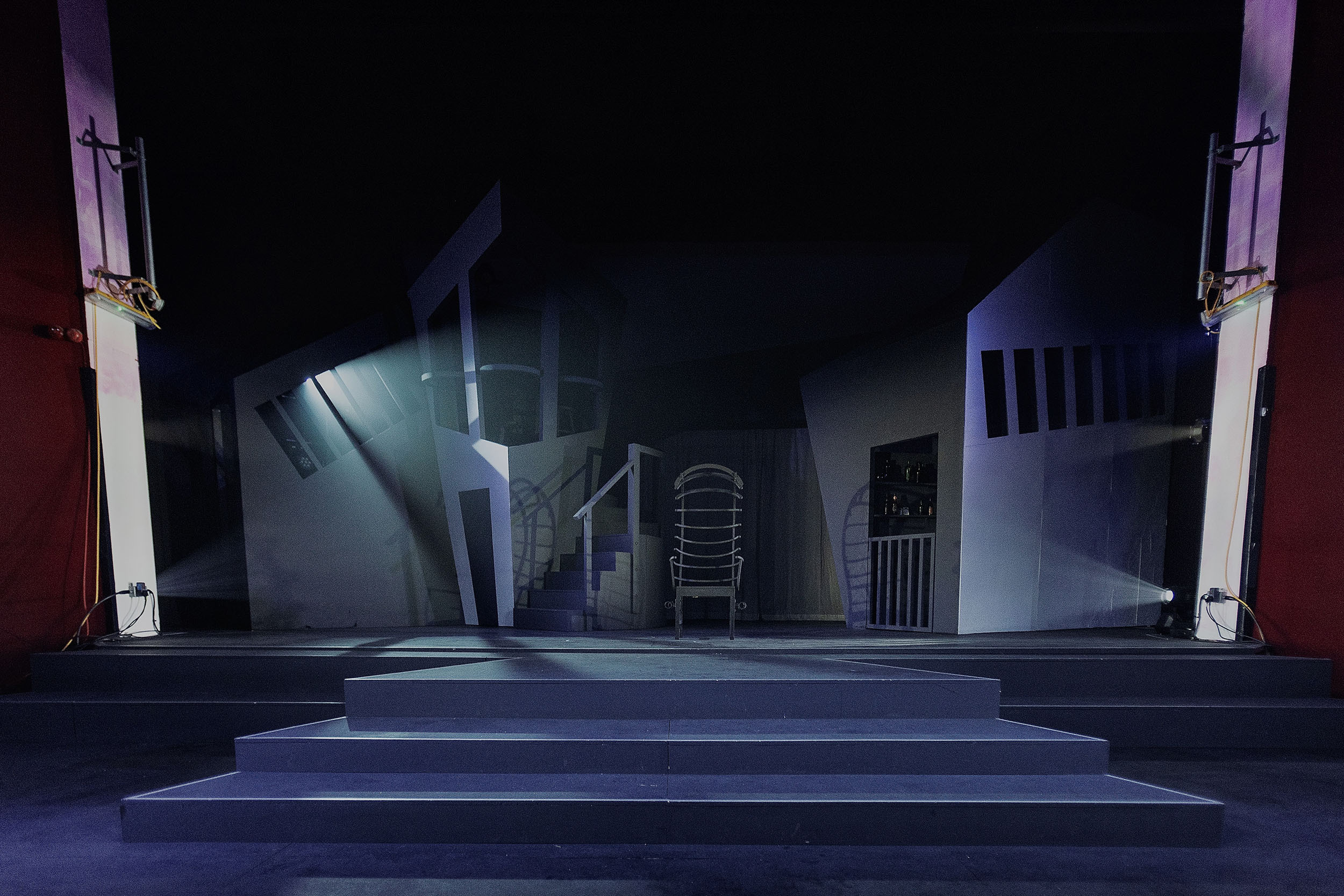 Theatre stage set with blue and purple lighting, white stairs centre stage, architectural elements including columns and railings in background.