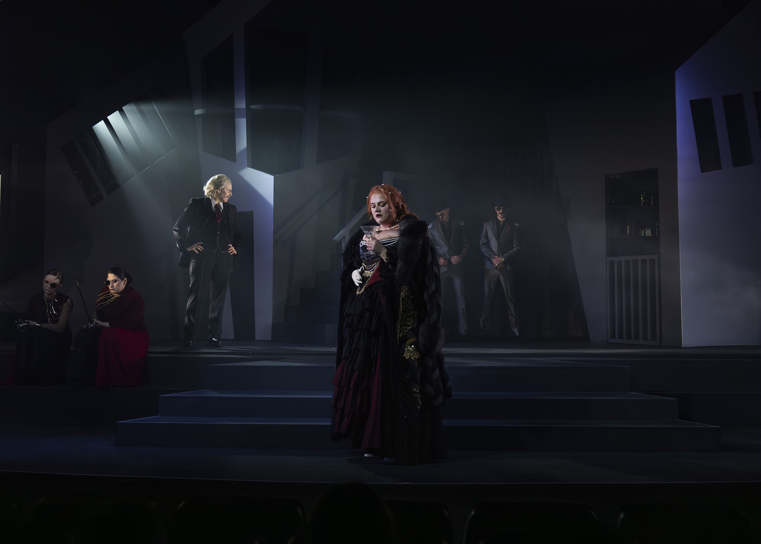 Theatre stage with dramatic lighting showing performers in dark costumes, blue-grey backdrop, bright spotlight from upper left.
