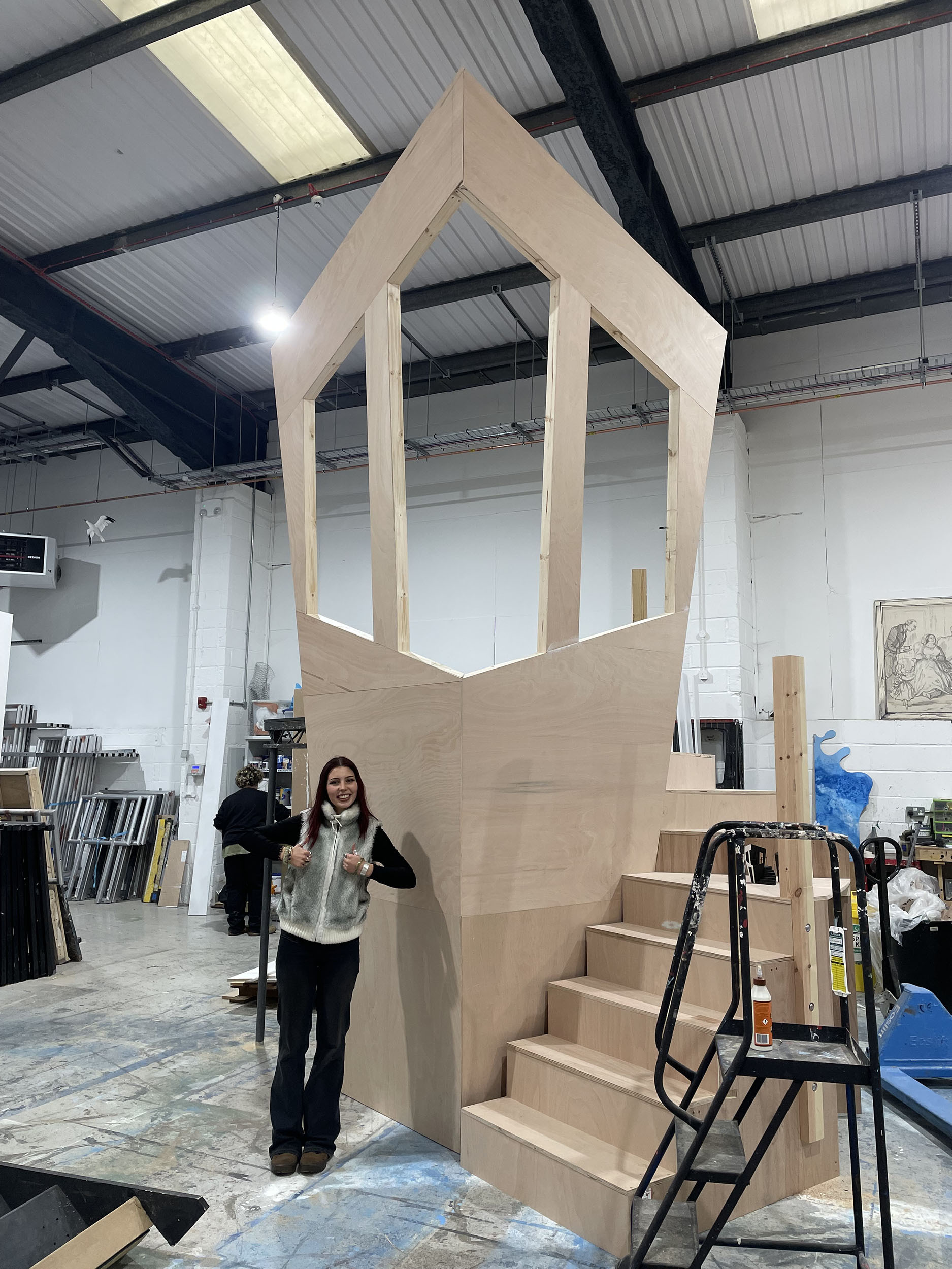Large wooden house-shaped structure with stairs in warehouse workshop. Woman stands beside pale timber construction under corrugated roof.