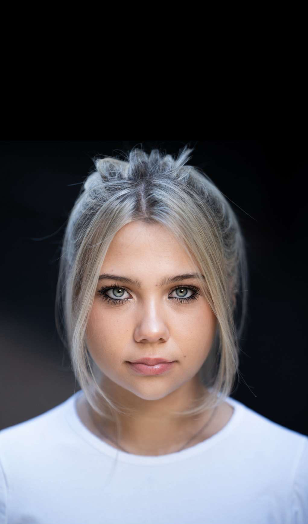 Young woman with platinum blonde hair in half-up style, wearing white top, looking directly at camera against dark background.