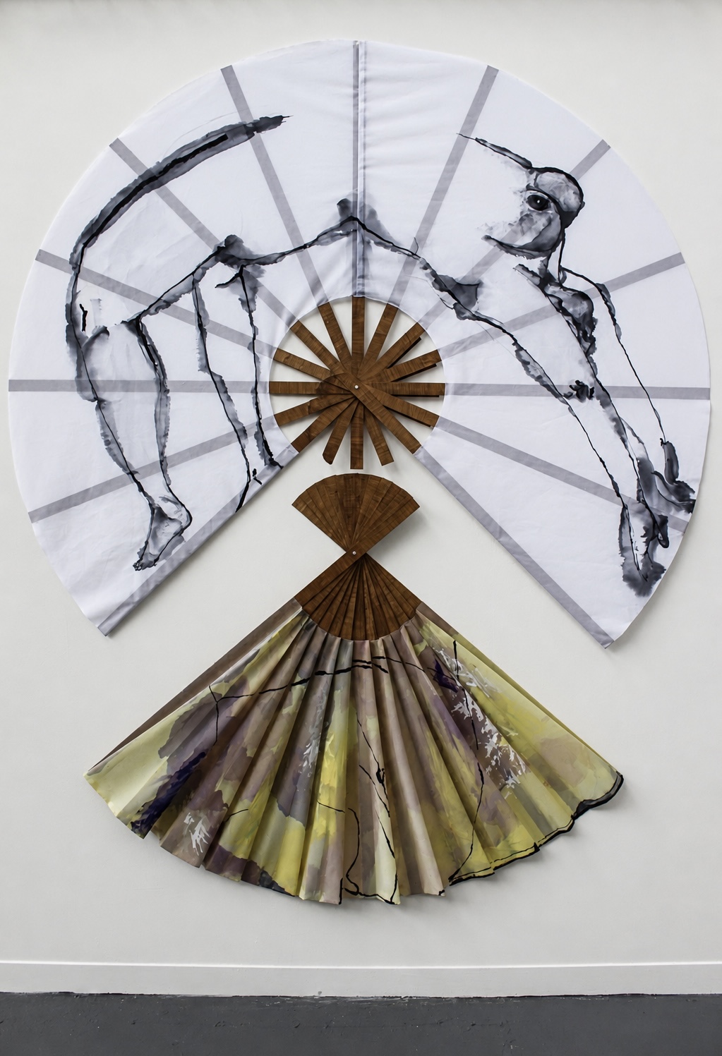 An artistic textile display featuring a large circular fabric with abstract black and white drawings, overlaid with a wooden fan shape. Below, a second fan-like fabric arrangement with a patterned design in earthy tones completes the composition.