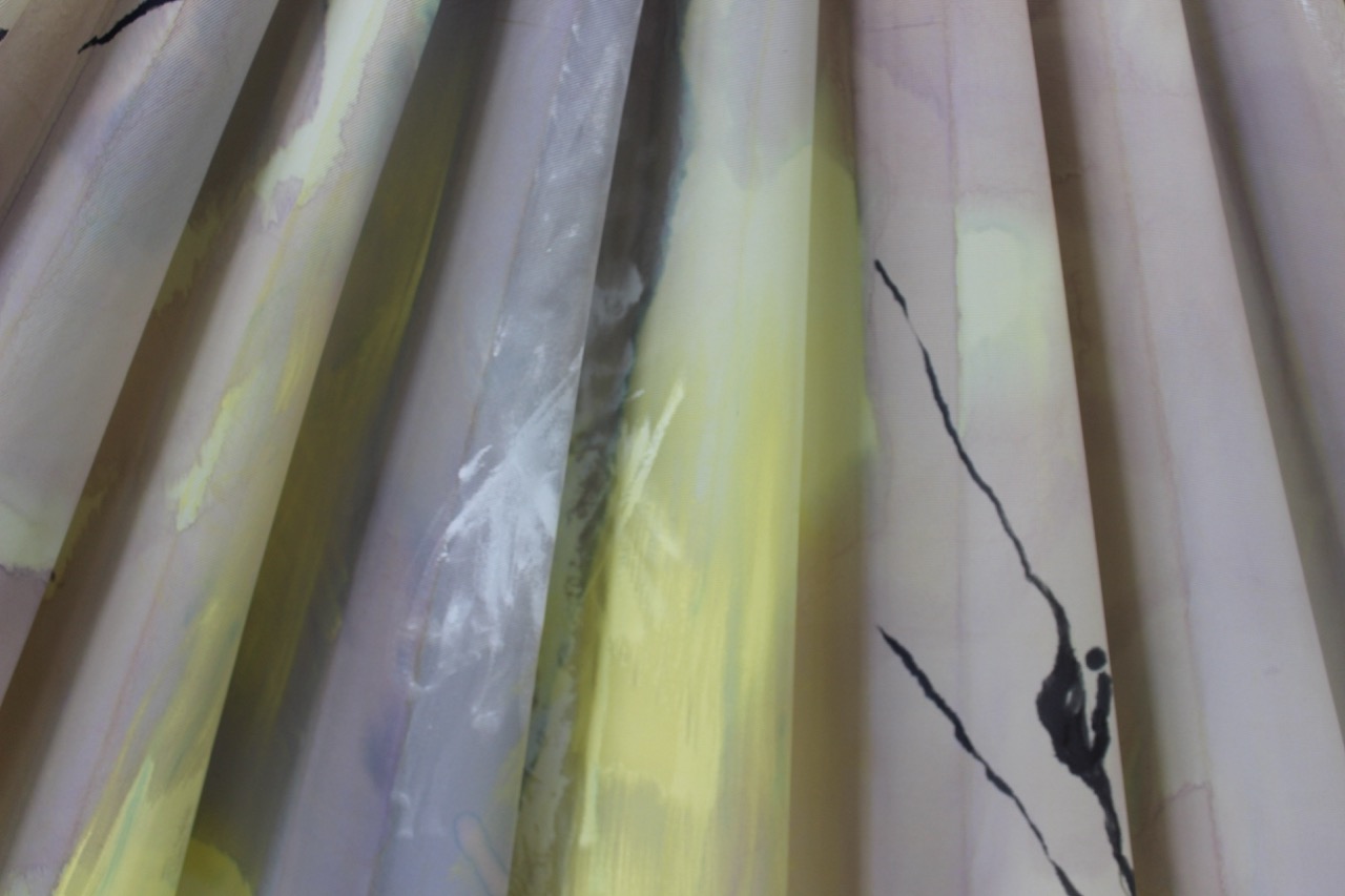 A close-up of folded fabric featuring soft pastel colours, including yellows and greys, with abstract black patterns. The fabric's texture appears smooth and flowing, highlighting its draped form.