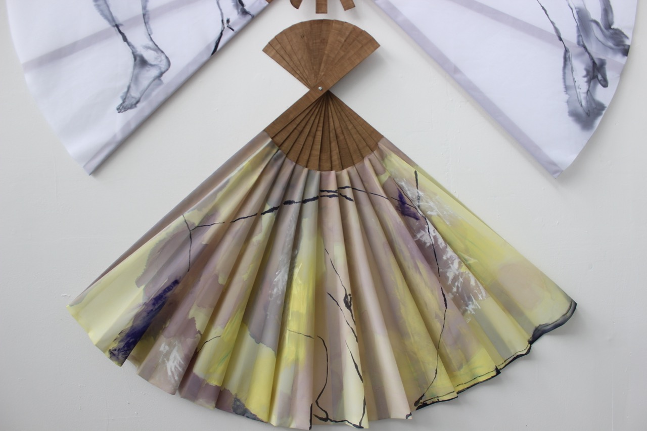 An artistic fan displayed on a white wall, featuring soft fabric in shades of yellow, purple, and beige, arranged in pleats. The fan's handle is made of wood with a curved, fan-shaped design. Two similar fans are positioned above it.