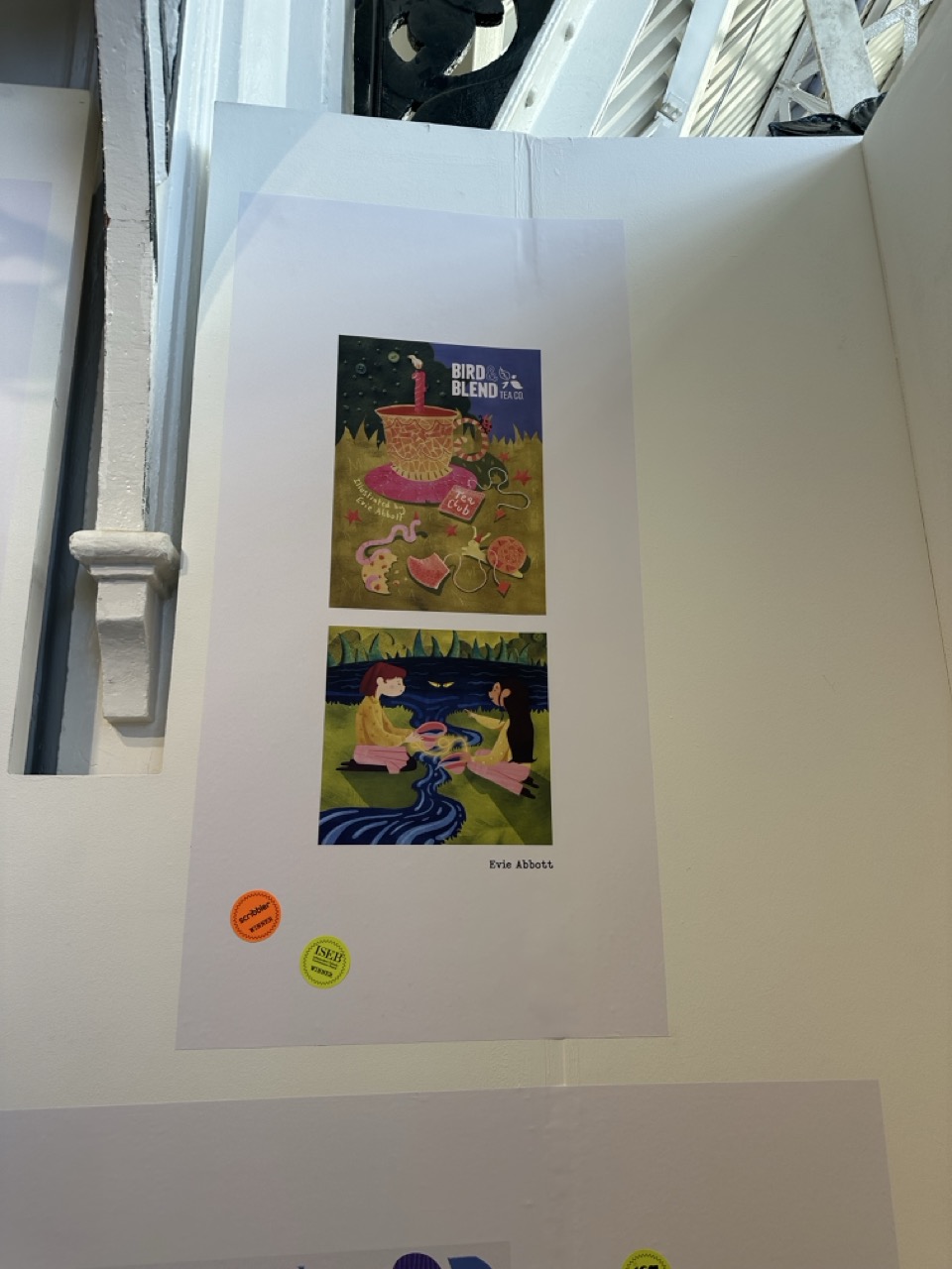 A poster displayed on a wall featuring two illustrated scenes. The top scene shows birds around a cake with the title 'Bird Blend'. The bottom scene depicts figures relaxing by a river in a green landscape. The artist's name, 'Erin Albrect', is noted at the bottom.