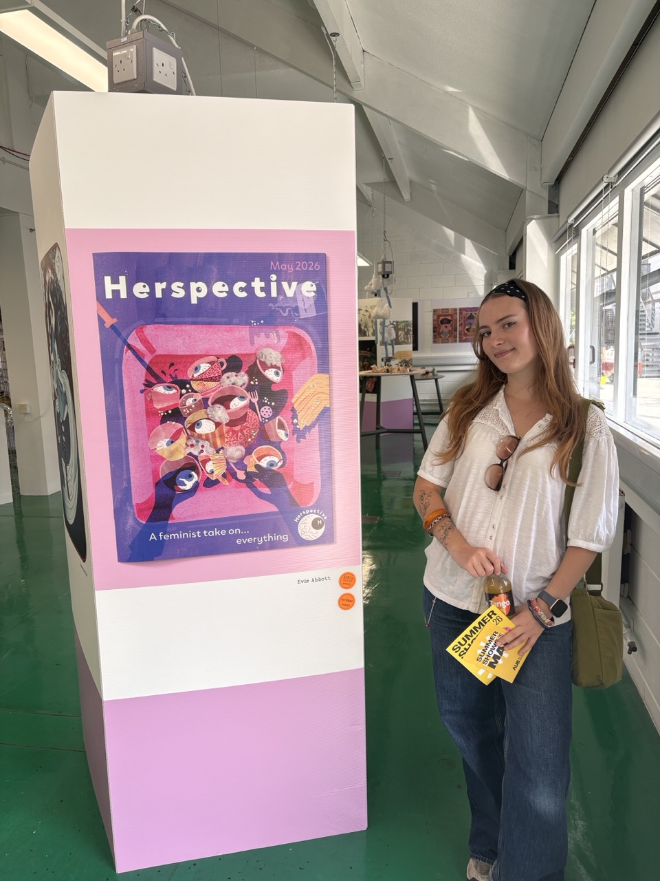 A young woman with long hair, wearing a white blouse and blue jeans, stands next to a tall white display featuring a colourful poster titled 'Herspective' showcasing various illustrations. The background includes art pieces and large windows.