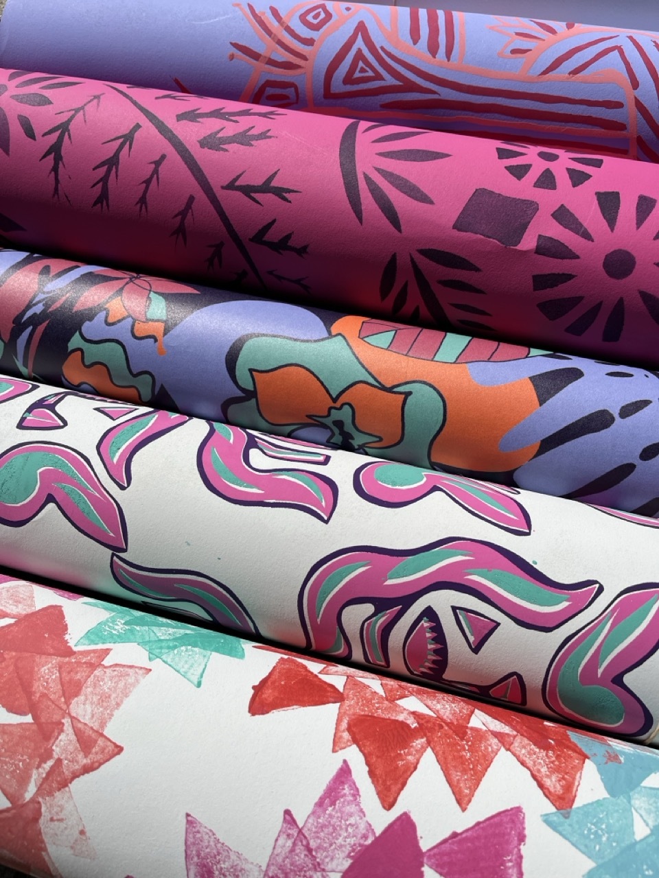 Stack of rolled fabric with colourful patterns: purple with red geometric designs, pink with black botanical motifs, multicoloured abstract shapes, white with pink swirls, and coral zigzag print.