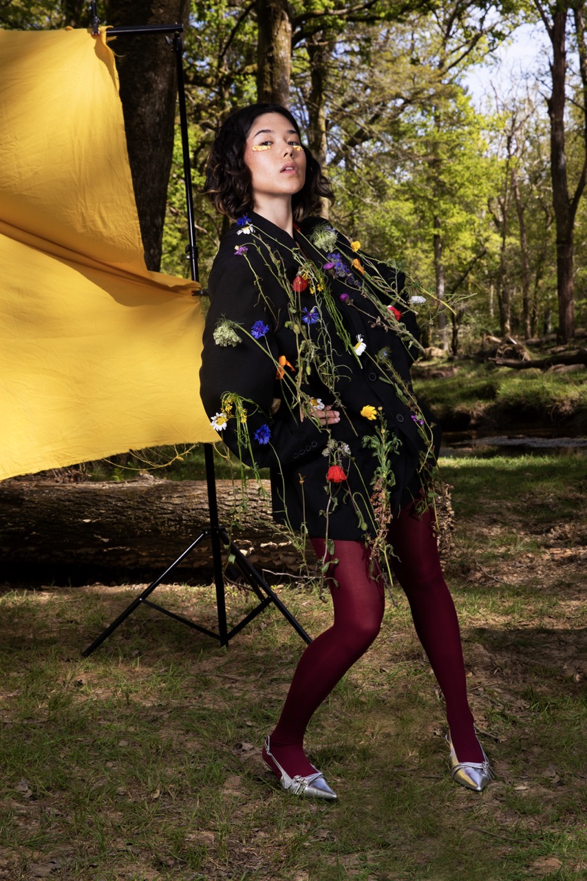 A woman stands in a forest wearing a black jacket adorned with floral decorations. She has curly hair and wears maroon tights, with silver shoes visible. In the background, there is a large yellow fabric draped between trees.