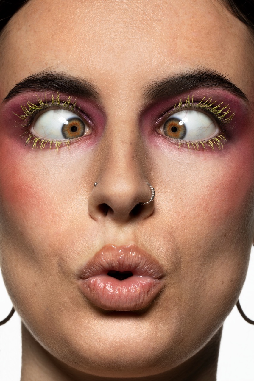 Close-up of a woman with dramatic makeup, featuring bold purple eyeshadow and yellow, spiky eyelashes. She has a nose ring and is pursing her lips, while her eyes have a striking, wide-open gaze, revealing brown irises against the expansive makeup.