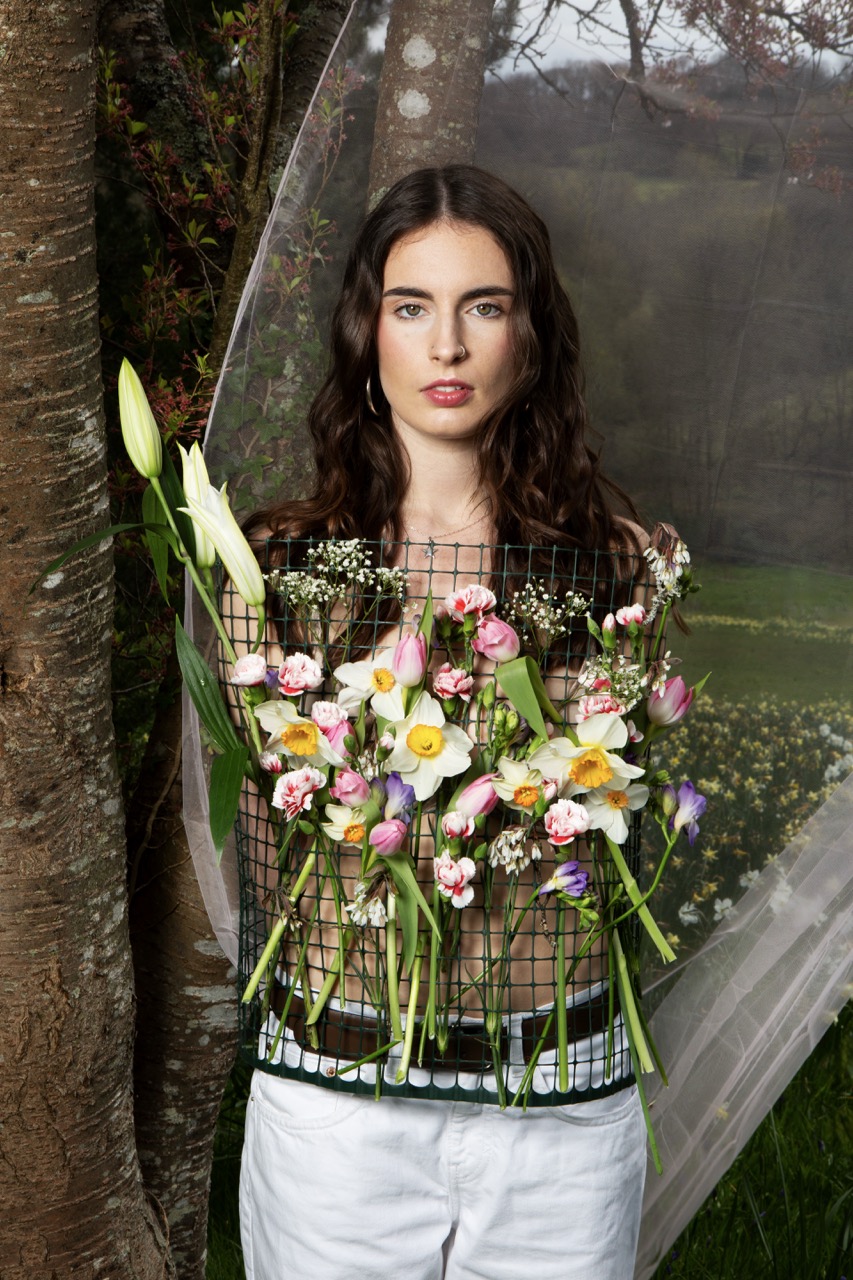 A woman with long brown hair stands against a backdrop of trees, holding a rectangular wire grid filled with various flowers, including lilies and daffodils. She wears a simple top and white trousers, with a sheer fabric draped in the background.