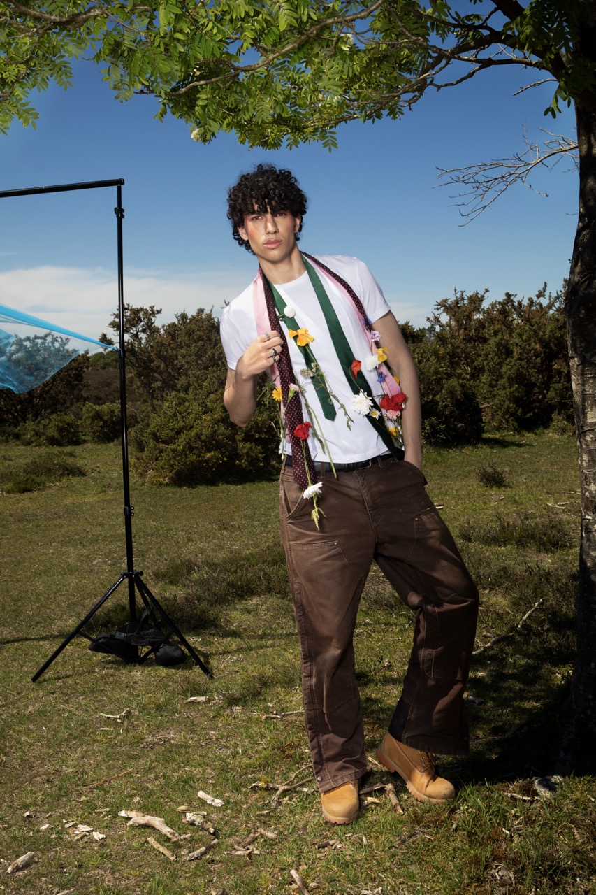 A young man with curly hair stands in a grassy field wearing a white t-shirt, brown trousers, and tan boots. He drapes a multicoloured scarf over one shoulder, with a backdrop of trees and a clear blue sky.