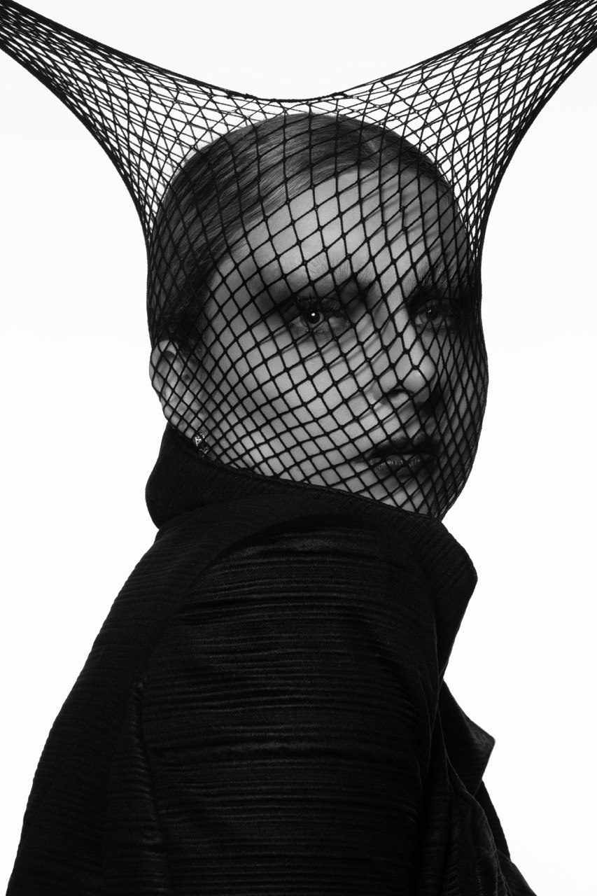A woman with short hair wears a black netted headpiece that creates a geometric shape above her head. She is dressed in a black jacket, and the image is presented in monochrome, highlighting the contrast between her features and the background.