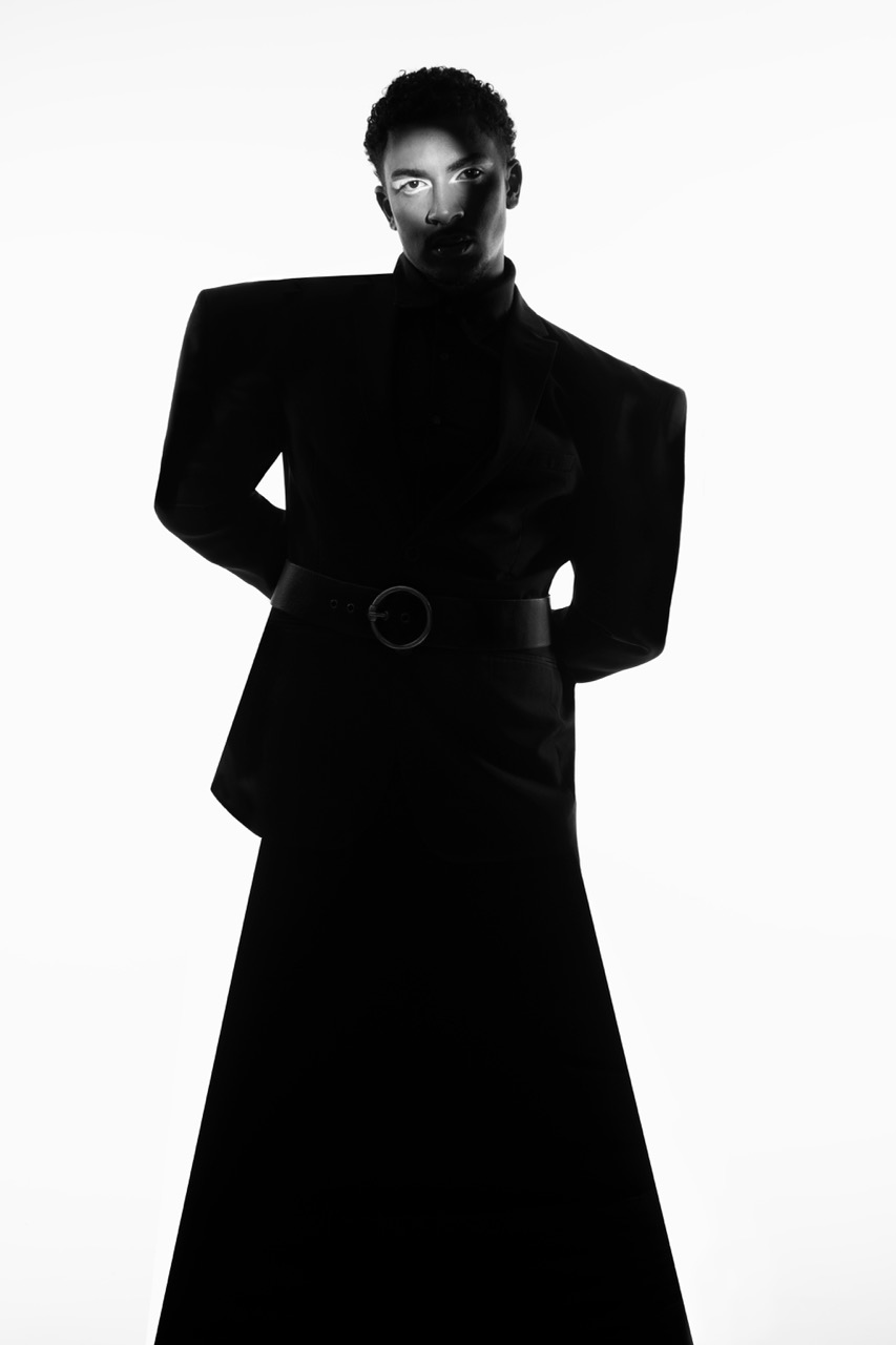 A model stands in silhouette wearing a structured black outfit with exaggerated shoulder pads and a long skirt. The model's hands are positioned behind their back, and the background is a stark white, enhancing the contrast of the garment.