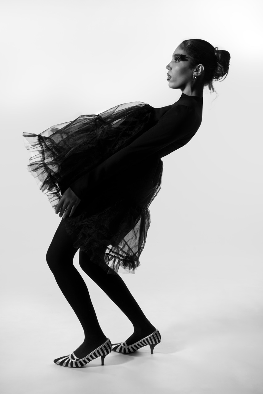 A model in a black dress with a voluminous tulle overlay poses in a dynamic stance. She wears black tights and black-and-white striped heels, with her hair pulled back and dramatic eye makeup, set against a white backdrop.