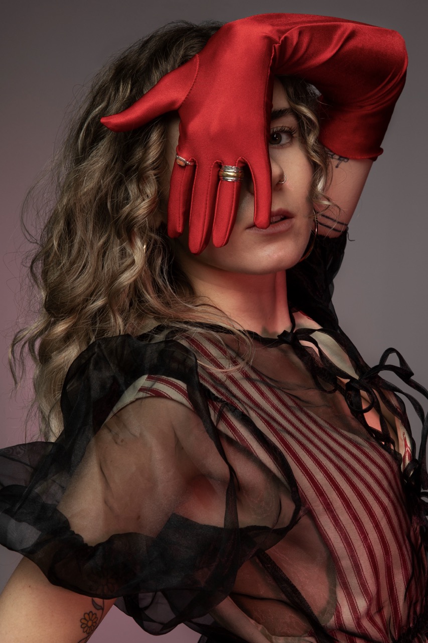 A woman with wavy hair wearing a black sheer top with ruffled sleeves poses while holding a red satin glove over her face. Gold rings adorn her fingers, and the background features a gradient of soft pink and grey tones.