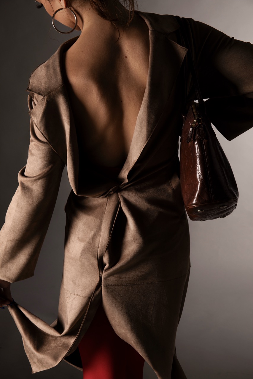 A person wearing a beige backless coat is seen from behind, holding the coat open with one hand. They are also carrying a brown handbag. The image features a neutral grey background, creating a focus on the outfit without any visible facial features.
