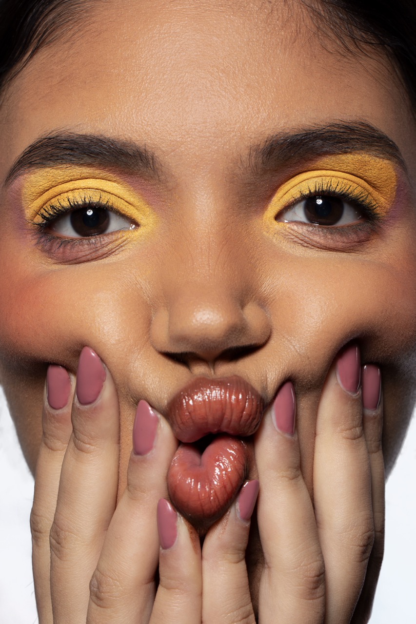 A close-up portrait of a young woman with yellow eyeshadow and peach-toned blush. She has her hands cupping her cheeks, creating a pouting expression with her lips, which are painted with a glossy shade. The background is softly blurred.