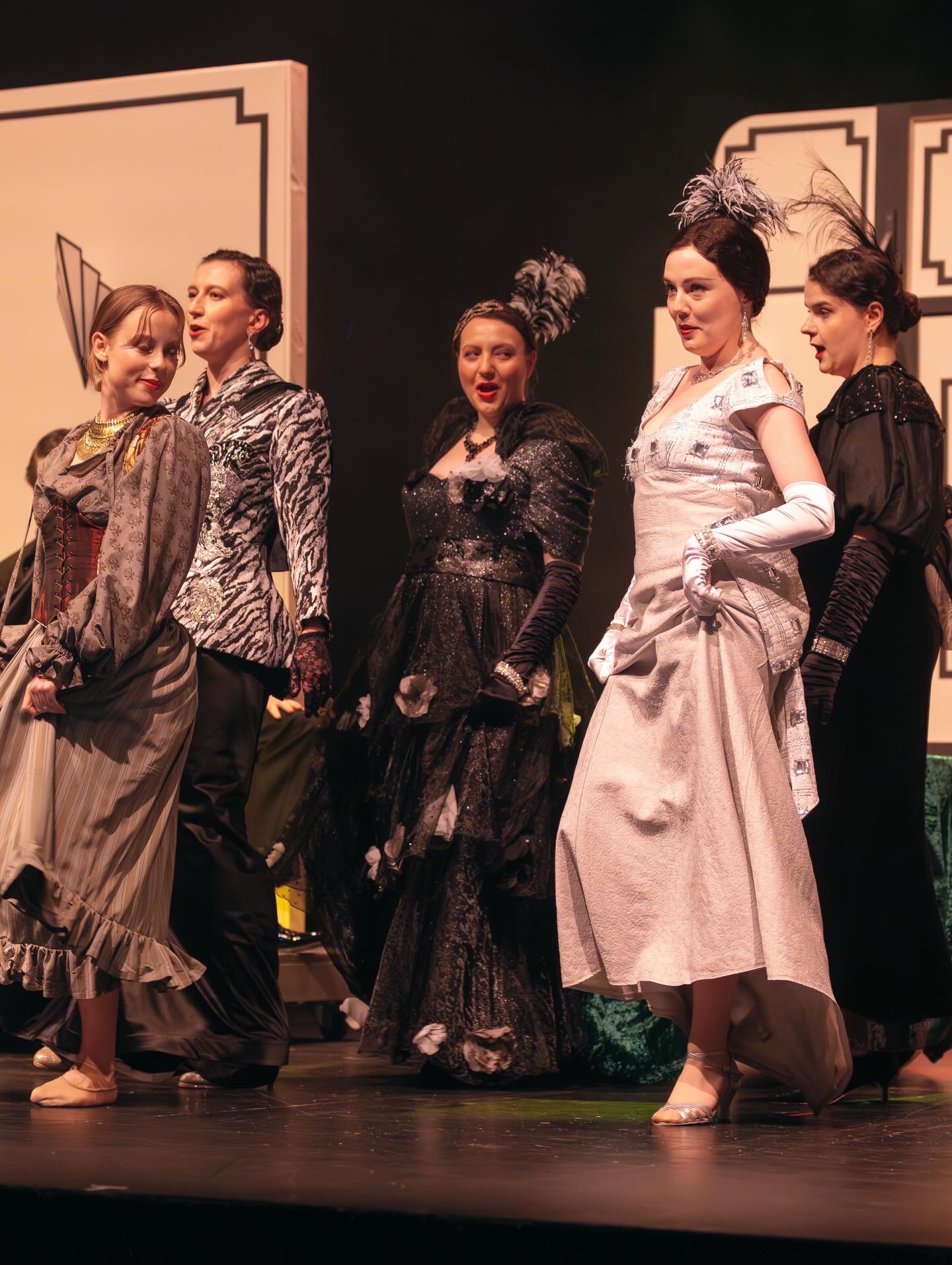 Five performers in historical costumes are seen on stage. They wear a variety of outfits, including a black dress with lace, a grey gown, and patterned jackets. The backdrop features art deco elements, enhancing the theatrical setting.