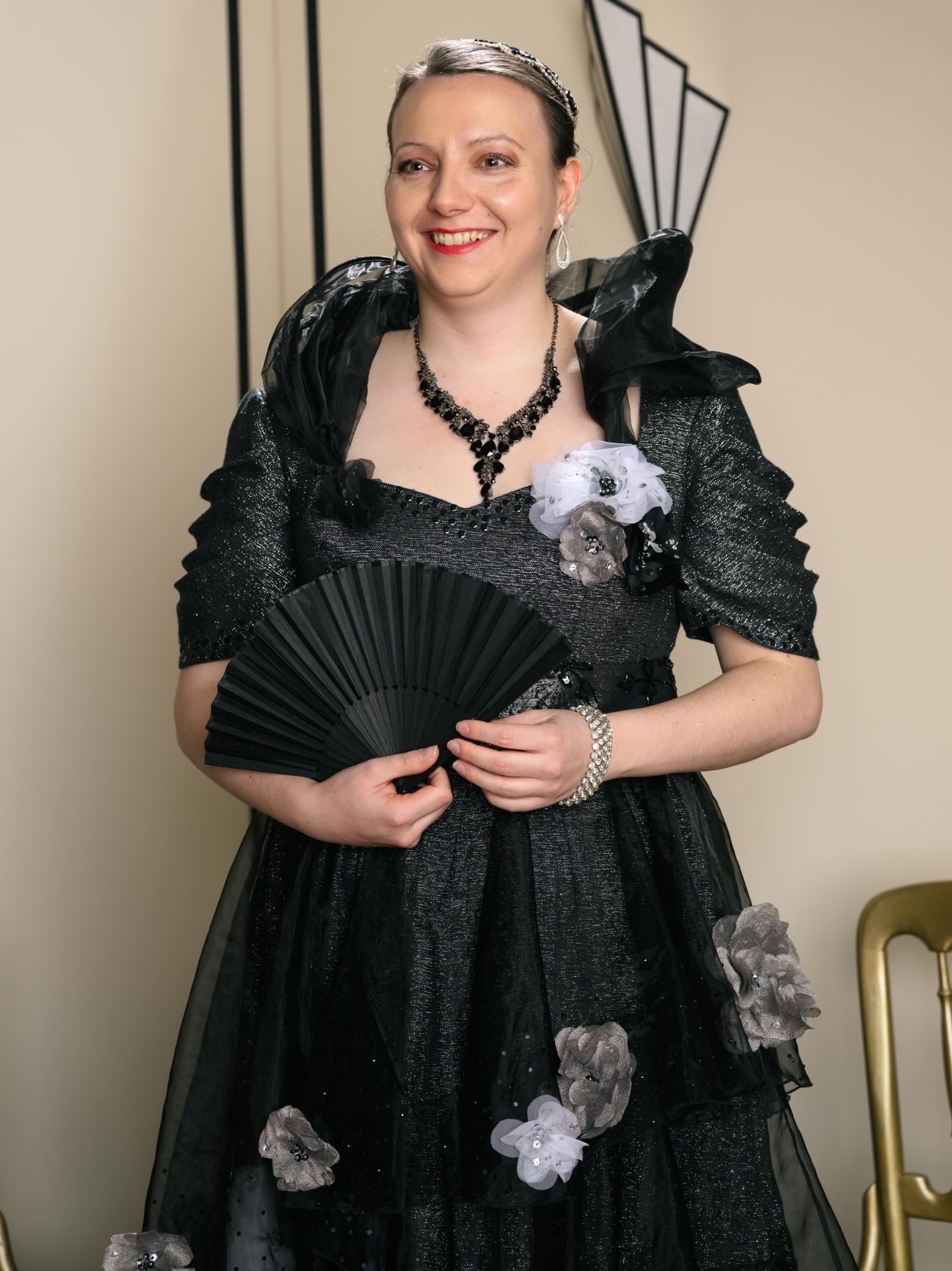 A woman in an elegant black dress adorned with fabric flowers poses while holding a black fan. She wears a necklace and has her hair styled with a decorative headpiece. The background features geometric shapes, adding a vintage flair.
