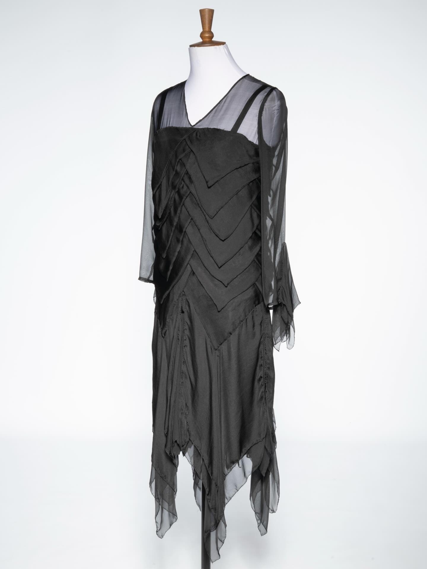 A black dress displayed on a mannequin. The dress features long sheer sleeves, a V-neck, and a chevron pattern across the bodice, with an asymmetric hem that is longer at the back, creating a flowing appearance.