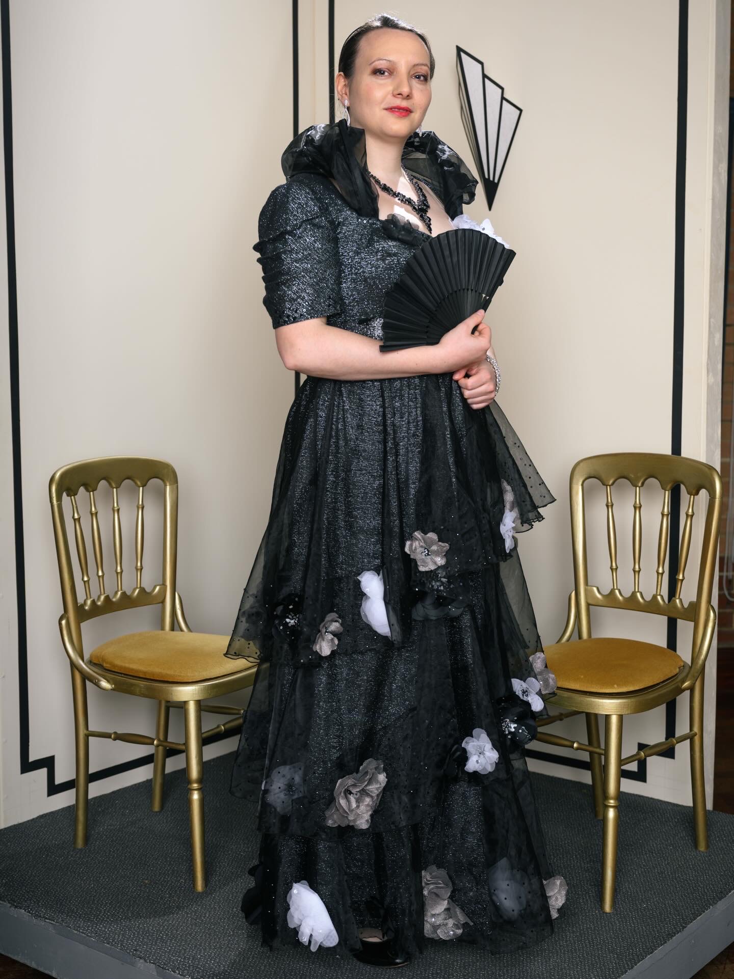 A woman in an elegant black gown adorned with floral decorations poses holding a black fan. Two gold chairs are visible on either side of her against a neutral backdrop.